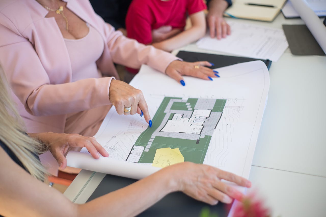 creative-03 A real estate agent in a pink blazer shows floor plans to clients, discussing property details.
